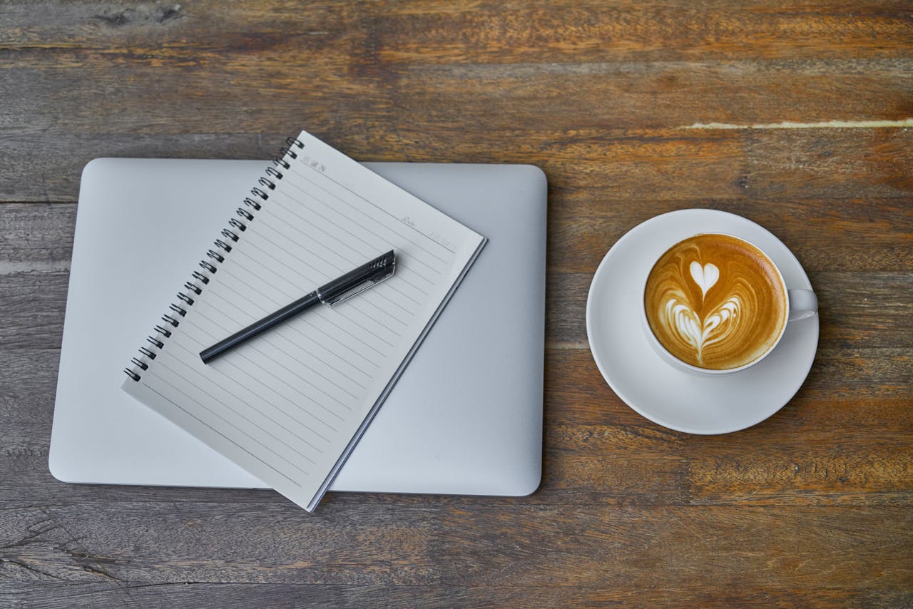 Flat lay of a coffee cup, notebook, pen, and laptop on a rustic wooden table.