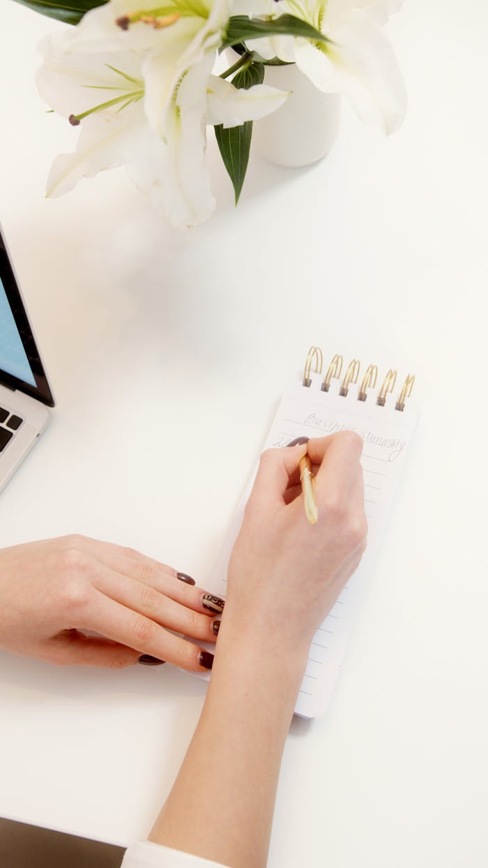 Female hand writing in notepad on a clean desk with flowers.